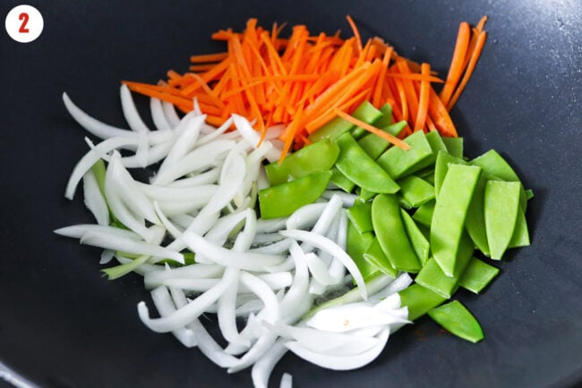 Sliced yellow onion, snow peas, and julienned carrots in a wok.