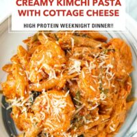 Closeup top view of plate with creamy kimchi pasta with shrimp topped with parmesan. Text overlay "Creamy Kimchi Pasta with Cottage Cheese", "High Protein Weeknight Dinner!!" and "That Spicy Chick".