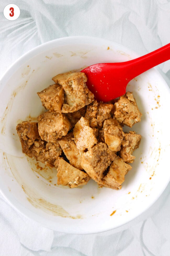 Bowl with torn tofu chunks coated in marinade and a red silicon spatula.