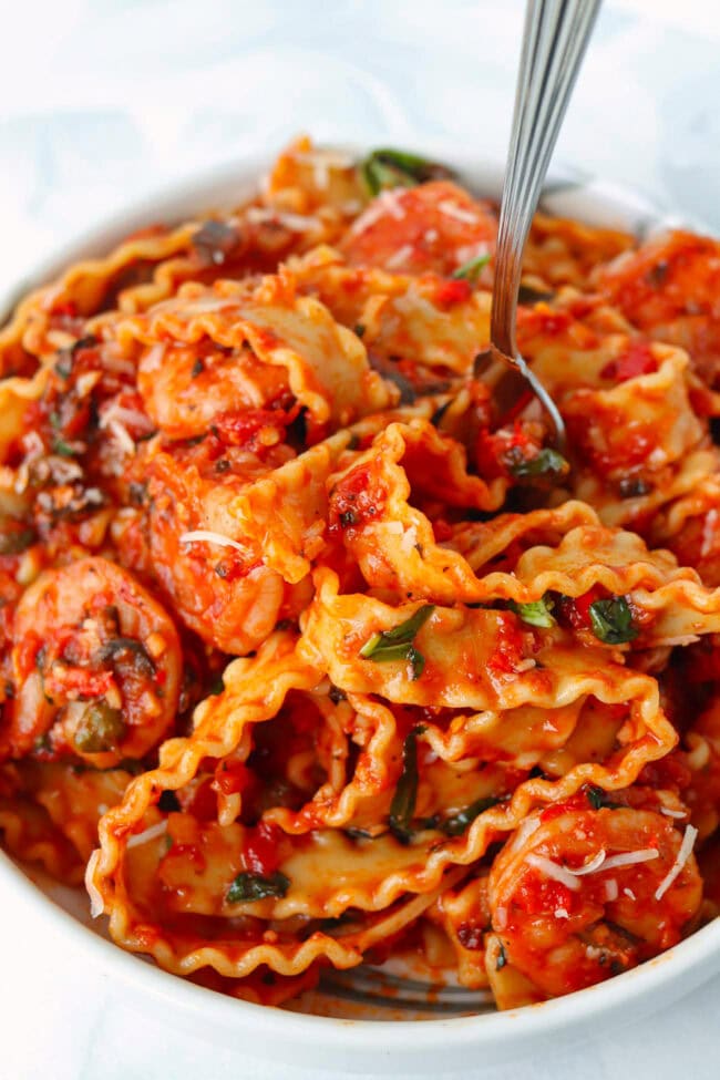 Closeup of mafaldine pasta coated in a spicy tomato sauce twirled around a fork in a plate with shrimp puttanesca.