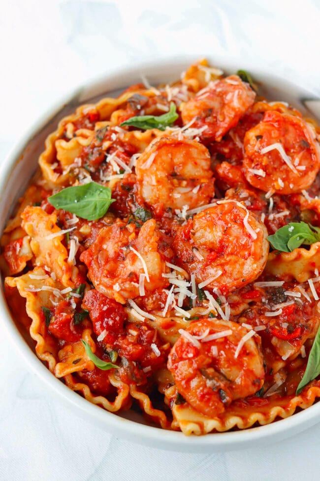 Closeup front view of plate with puttanesca pasta with shrimp garnished with grated cheese and basil leaves.