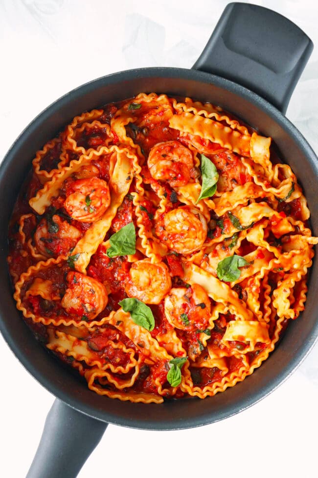 Puttanesca pasta with shrimp garnished with basil leaves in a skillet.
