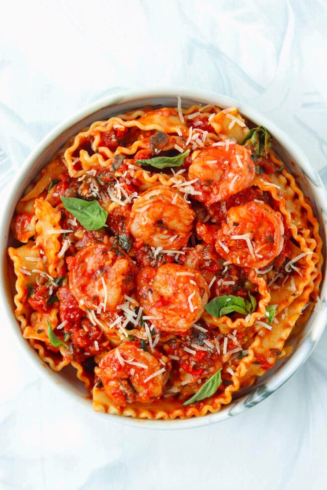 Plate with puttanesca pasta with shrimp garnished with grated cheese and basil leaves.