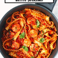 Skillet with spicy tomato sauce pasta with shrimp topped with basil leaves. Text overlay "Pasta Puttanesca with Shrimp" and "That Spicy Chick".