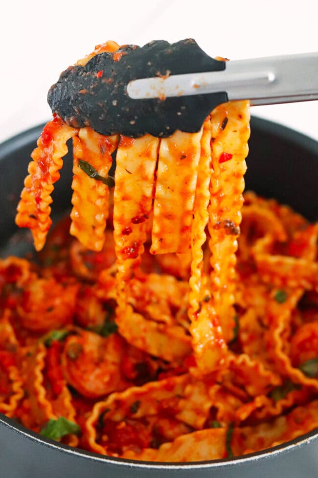 Tongs holding up mafaldine pasta coated in a spicy tomato sauce above a pan.