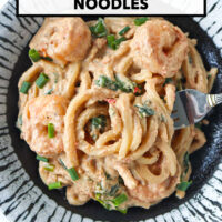 Fork in a plate with creamy tahini udon noodles with shrimp, spinach and green onions. Text overlay "Spicy Tahini Peanut Noodles" and "That Spicy Chick".