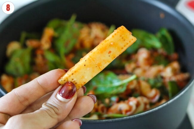 Hand holding a Parmigiano Reggiano cheese rind above a pan with tomato basil pasta.