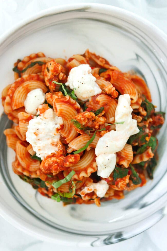 Closeup of plate with tomato basil pasta topped with torn burrata that's seasoned with black pepper.