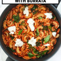 Skillet of tomato pasta with spinach topped with basil and black pepper seasoned torn burrata. Text overlay "Tomato Basil Pasta with Burrata" and "That Spicy Chick".